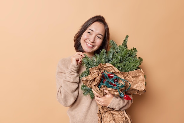 Die horizontale Aufnahme einer verträumten asiatischen Frau schaut glücklich weg, mit zufriedenem Ausdruck, hält grüne Zweige, die in einem Blumenstrauß angeordnet sind, und eine Retro-Girlande trägt einen lässigen Pullover, der isoliert über beigem Hintergrund liegt