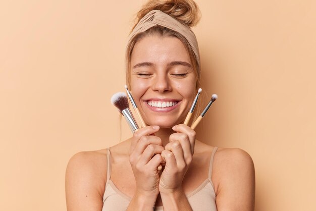 Die horizontale Aufnahme einer zufriedenen Frau schließt die Augen, lächelt breit und hält Kosmetikpinsel zum Auftragen von Make-up trägt ein lässiges T-Shirt mit Stirnband, das über beigem Hintergrund isoliert ist. Gesichtspflegekonzept.