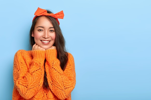 Die horizontale Aufnahme einer zufriedenen schönen asiatischen Dame berührt das Kinn mit beiden Händen, lächelt angenehm, hat langes dunkles Haar und trägt einen orangefarbenen Pullover