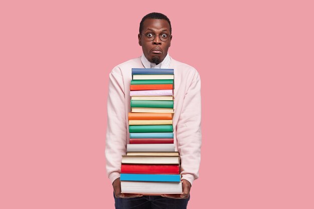 Die horizontale Aufnahme eines afroamerikanischen Studenten hat einen verwirrten Ausdruck, einen überraschten Blick, eine dicke Enzyklopädie und wissenschaftliche Literatur