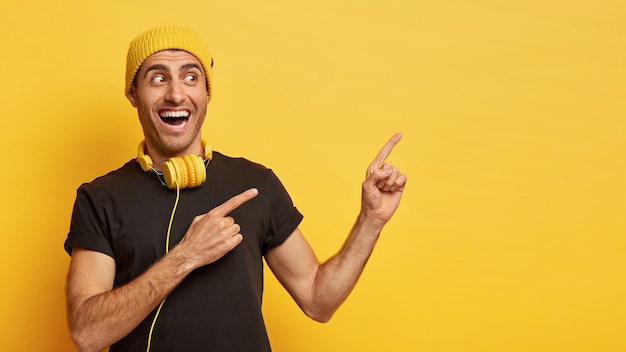 Die horizontale Aufnahme eines glücklichen europäischen Mannes zeigt mit zwei Zeigefingern, gekleidet in stilvolle schwarze und gelbe Kleidung, und trägt moderne Kopfhörer um den Hals, um ein Lied zu hören