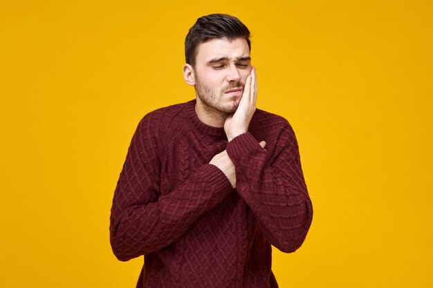 Die horizontale Aufnahme eines glücklichen, frustrierten jungen Mannes in einem gestrickten Pullover, der Probleme mit der Zahnhöhle hat, muss den Zahnarzt sehen, der die Hand auf der Wange hält und das Gesicht verzieht. Er kann keine schrecklichen Zahnschmerzen ertragen