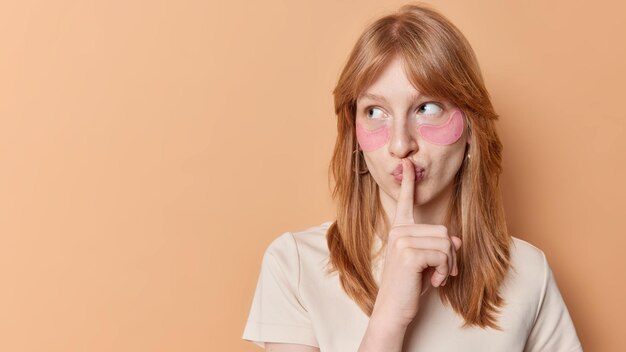 Die horizontale Aufnahme eines Ingwermädchens trägt Schönheitsflecken unter den Augen auf, um Schwellungen zu entfernen, die mysteriös aussehen, gekleidet in einem lässigen T-Shirt, isoliert über beigem Hintergrund, leerer Raum für Werbung