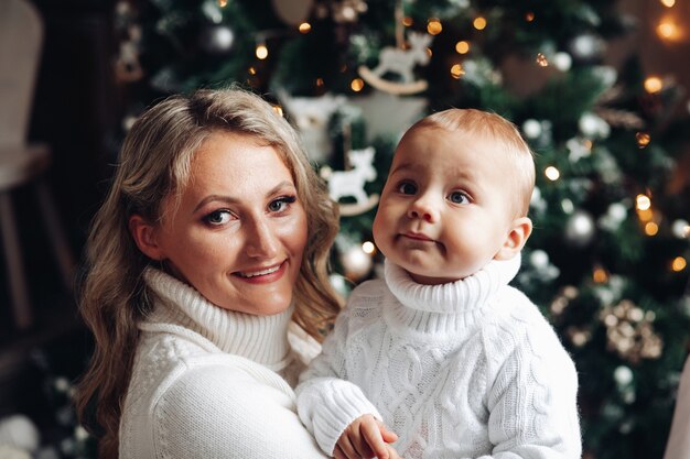 Die hübsche Mutter hat viel Spaß mit ihrem Baby in der Nähe des Weihnachtsbaumes zu Hause und lächelt