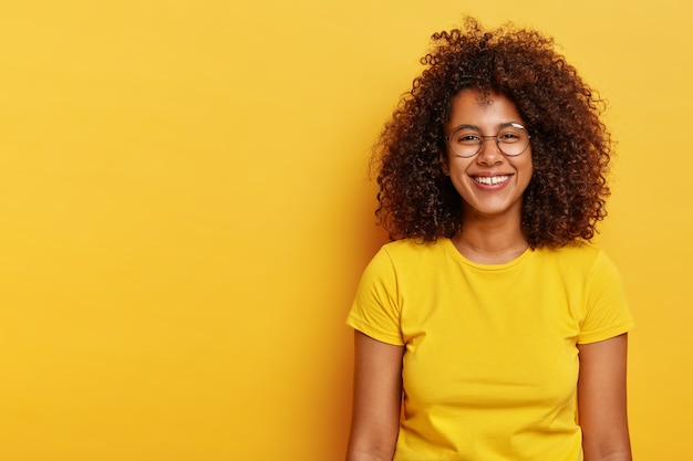 Die isolierte Aufnahme eines glücklichen hübschen Afro-Mädchens hat buschiges dunkles Haar, trägt eine große runde Brille, ein leuchtend gelbes T-Shirt, lächelt glücklich, ist froh, einen erfolgreichen Tag zu haben, modelliert drinnen, fühlt sich entspannt und sorglos