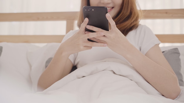 Die junge Asiatin, die Smartphone verwendet, beim morgens liegen auf Bett nach aufwachen