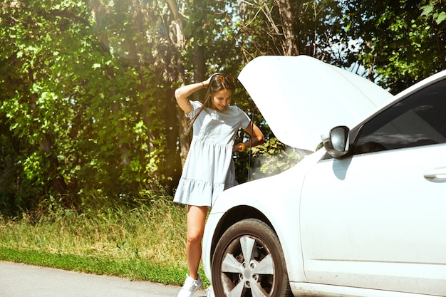 Die junge Frau brach auf dem Weg zur Ruhe das Auto zusammen. Sie versucht, das kaputte selbst zu reparieren oder sollte per Anhalter fahren. Nervös werden. Wochenende, Probleme auf der Straße, Urlaub.