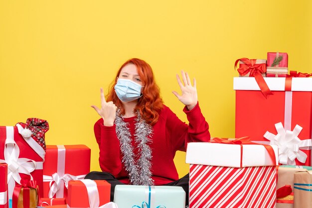 Die junge Frau der Vorderansicht, die um Weihnachten sitzt, präsentiert in der Maske auf gelbem Hintergrund Neujahrsvirus-Emotions-Covid-Farbe