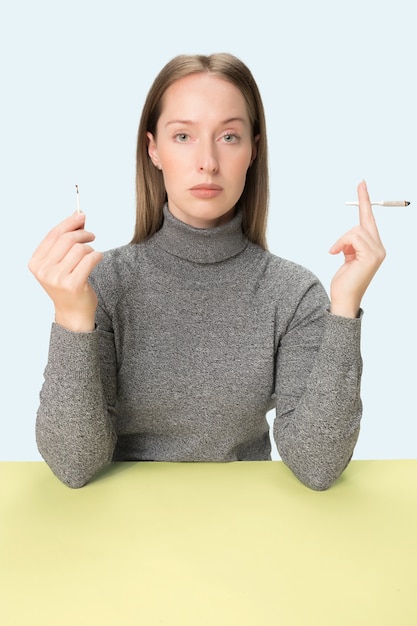 Die junge Frau, die Zigarette raucht, während am Tisch im Studio sitzt. Trendige Farben. Das Porträt des kaukasischen Mädchens im Minimalismusstil mit Kopierraum