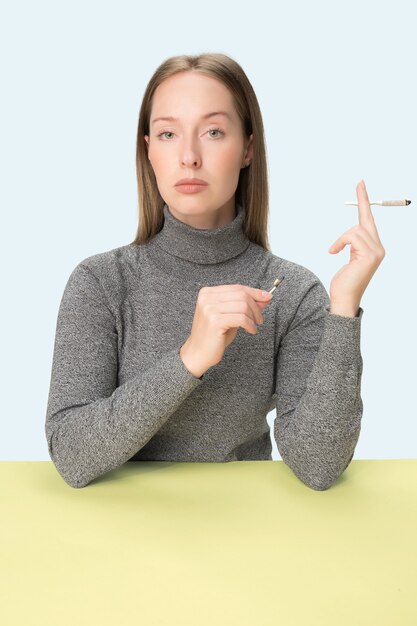 Die junge Frau, die Zigarette raucht, während sie am Tisch im Studio sitzt. Trendige Farben. Das Porträt des kaukasischen Mädchens im Minimalismusstil mit Kopienraum