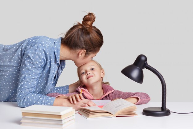 Die junge Frau kümmert sich um ihr Kind, küsst die Tochter auf die Stirn, lobt sie für das gute Lernen, erklärt Material, liest Bücher und bereitet sich auf den Schulunterricht vor, isoliert auf Weiß. Studienkonzept