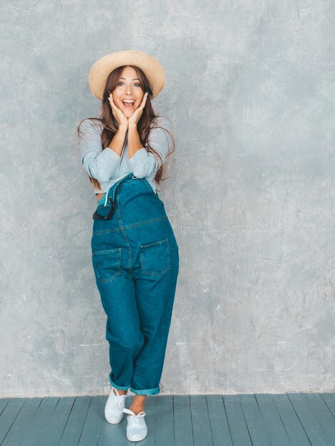 Die junge schöne überraschte Frau, die mit den Händen schaut, nähern sich Gesicht. Modisches Mädchen in der zufälligen Sommeroverallkleidung und -hut. Frau, die nahe grauer Wand im Studio aufwirft