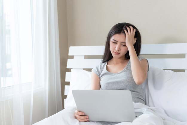 Die junge Schönheit, die auf Bett sitzt, sie benutzt das ernsthafte Laptopgefühl und die Kopfschmerzen