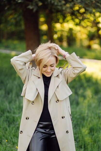 Die kaukasische blonde Frau trägt ein Grabenlächeln glücklich am sonnigen Frühlingstag draußen, der im Park geht