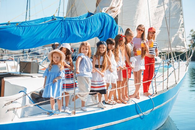 Die Kinder an Bord der Seelyacht trinken Orangensaft. Die Teenager- oder Kindermädchen gegen blauen Himmel im Freien. Farbenfrohe Kleider. Kindermode, sonnige Sommer-, Fluss- und Ferienkonzepte.