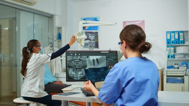 Die Krankenschwester des Stomatologen vergleicht Röntgenaufnahmen mit Blick auf den Computer, während der Facharzt mit Gesichtsmaske mit einem Mann mit Zahnschmerzen spricht, der auf einem stomatologischen Stuhl sitzt und Werkzeuge für die Operation vorbereitet