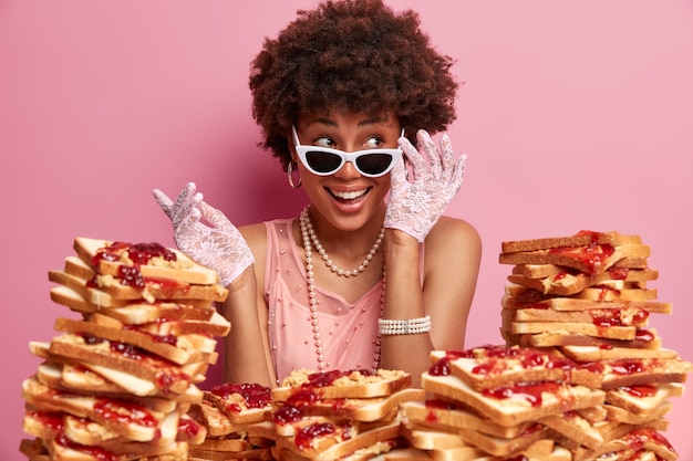 Kostenloses Foto die lächelnde ethnische dame schaut zur seite und hält die hand auf der sonnenbrille, ist gut gelaunt, kichert positiv auf der party, trägt stilvolle kleidung, posiert über einer rosigen wand, viele leckere sandwiches herum