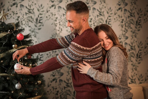 Die lächelnde Frau, die Mann von der Rückseite in den Strickjacken umarmt, nähern sich Weihnachtsbaum