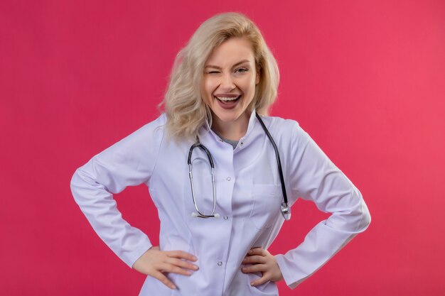 Kostenloses Foto die lächelnde junge ärztin, die ein stethoskop im medizinischen gewand trug, legte ihre hände auf die hüften des roten hintergrunds