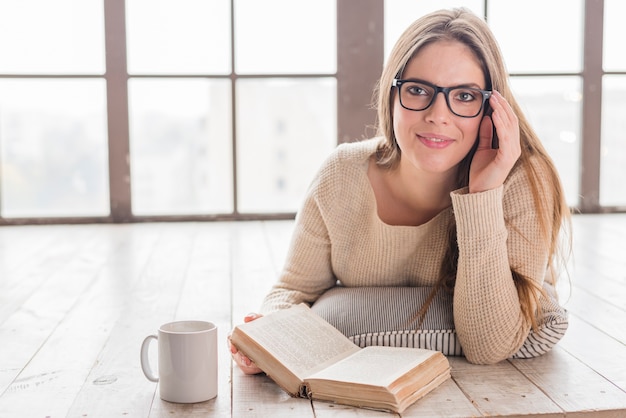 Die lächelnde junge Frau, die auf Massivholzboden mit liegt, überreichen die Brillen, die Buch halten
