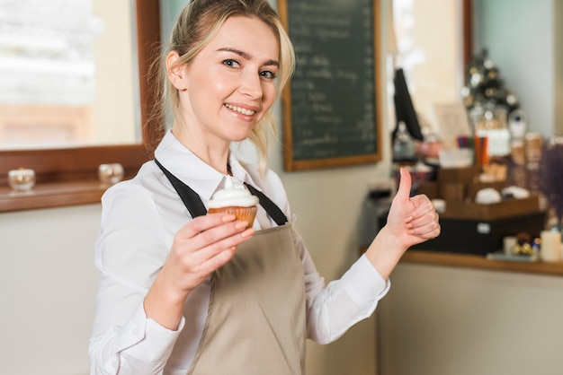 Die lächelnde junge Frau, die in der Hand gebackene Muffins zeigt, zeigt Daumen herauf Zeichen