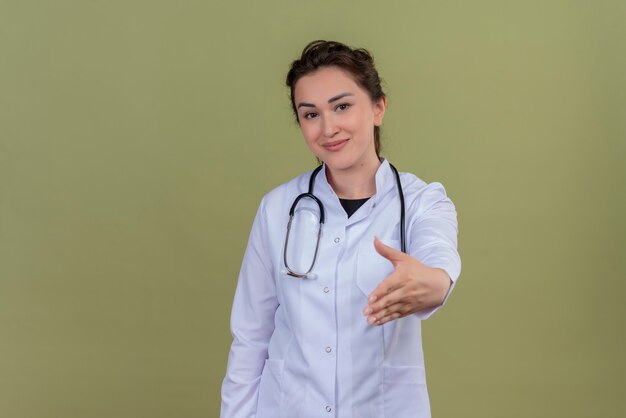 Die lächelnde junge Ärztin im medizinischen Kleid mit dem Stethoskop streckte ihre Hand an der grünen Wand aus