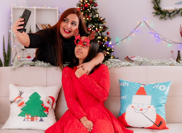 Die lächelnde Mutter nimmt Selfie mit ihrer Tochter in der Rentiermaske, die auf Couch sitzt und Weihnachtszeit zu Hause genießt