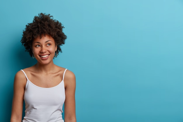 Die lächelnde positive junge Frau mit Afro-Haaren schaut mit verträumtem Gesichtsausdruck weg, denkt über eine gute Zukunft nach, hat ein zahniges Lächeln, trägt ein lässiges T-Shirt, posiert gegen die blaue Wand, leerer Raum für Text