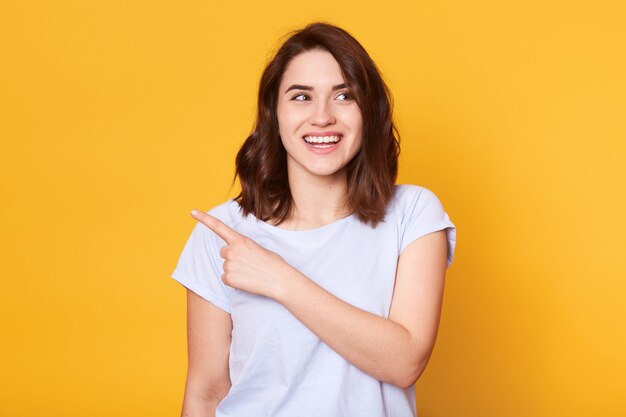 Die lächelnde schöne junge Frau trägt ein weißes Freizeithemd, zeigt mit dem Zeigefinger zur Seite