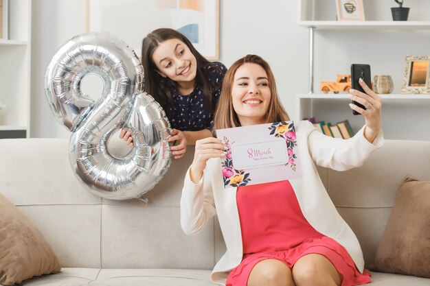 Die lächelnde Tochter, die hinter dem Sofa steht und die Ballonmutter mit der Nummer acht hält, mit der Grußkarte auf dem Sofa sitzend, macht ein Selfie am glücklichen Frauentag im Wohnzimmer