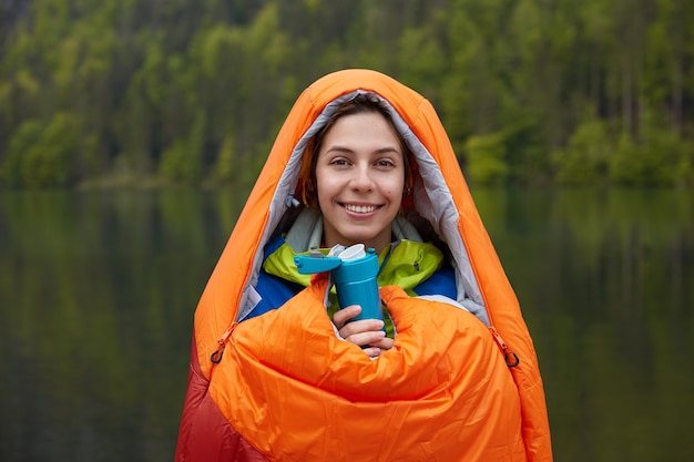 Die lächelnde weibliche reisende ist gut gelaunt, in einen schlafsack gewickelt, verbringt ihre freizeit in der natur und hält eine thermoskanne mit heißem getränk