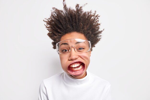 Die lustige junge Afroamerikanerin hat einen weit geöffneten Mund, trägt eine transparente Brille, schreit laut.