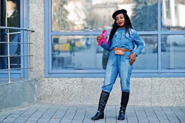 Die Macht zu kämpfen Stilvolle, modische afroamerikanische Frauen in Jeans und schwarzer Baskenmütze gegen moderne Gebäude mit rosa Schleife für Brustkrebs
