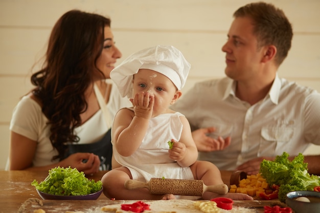 Die mutter, der vater und das kleine kind sitzen auf dem tisch in der nähe von teig und gemüse