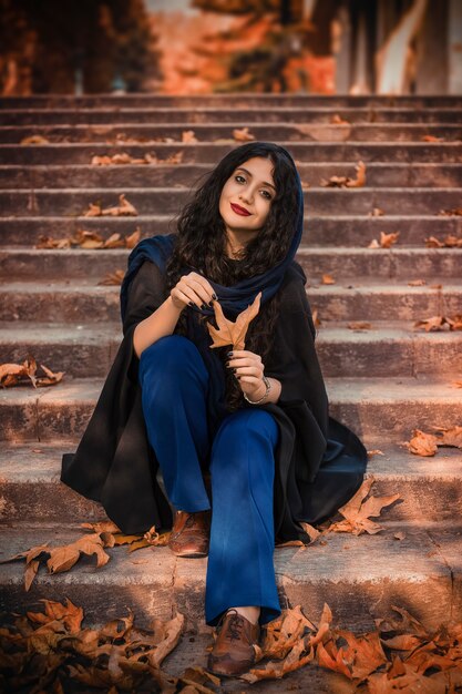 Die reizende junge Frau trägt einen gestrickten Schal und ein schwarzes Kleid und sitzt auf der Treppe im Herbstpark.