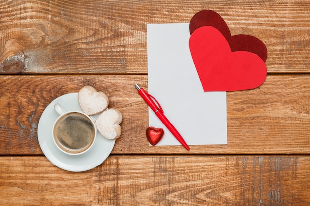 Die roten Herzen und das leere Blatt Papier und Stift auf hölzernem Hintergrund mit einer Tasse Kaffee