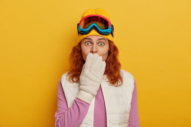 Die schockierte rothaarige Frau bedeckt den Mund und starrt in die Kamera, trägt spezielle Kleidung und Ausrüstung für den Wintersport, fährt gerne Ski oder Snowboard.