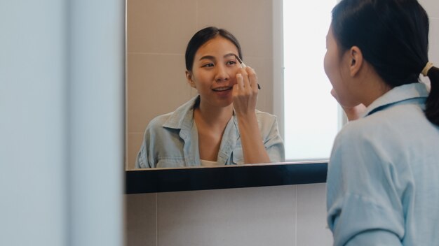 Die schöne Asiatin, die Augenbrauenstift verwendet, bilden im vorderen Spiegel, die glückliche lateinische Frau, die Schönheitskosmetik verwendet, um sich zu verbessern, die zum im Badezimmer zu Hause arbeiten bereit ist. Lifestyle-Frauen entspannen zu Hause.