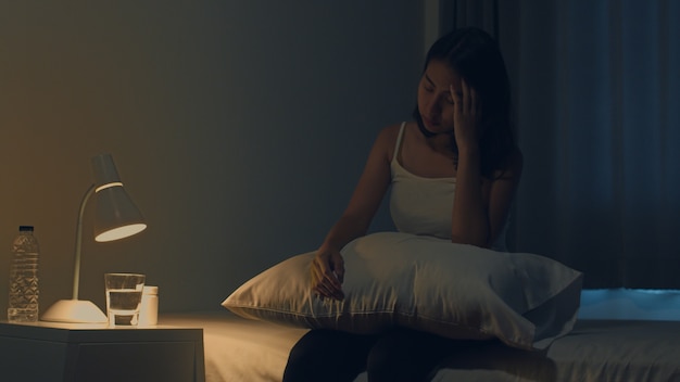 Die schöne asiatische junge Frau, die auf Bett sitzt, nehmen Schlafenpille oder Nachtmedizin im Schlafzimmer. Ungesunde kranke Inderin leidet an Schlaflosigkeit oder Kopfschmerzen, depressives Mädchen hält Antidepressiva.