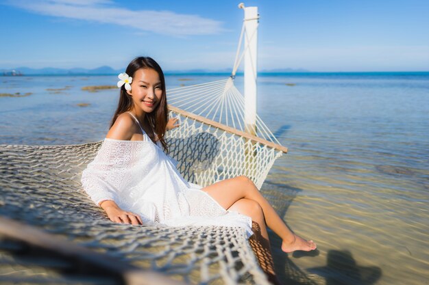 Die schöne junge asiatische Frau des Porträts, die auf Hängematte um Seestrandozean für sitzt, entspannen sich