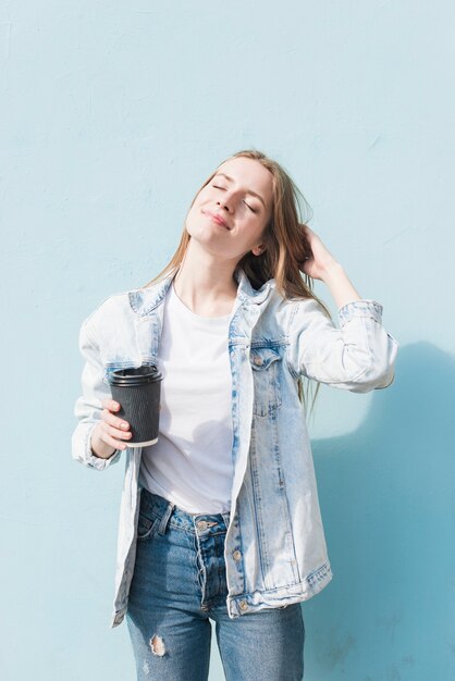 Die schöne junge Frau, die Wegwerfkaffeetasse mit Augen hält, schloss stehende nahe Wand