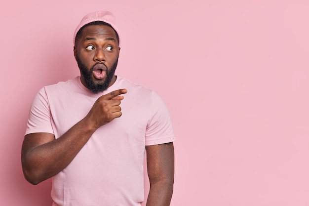 Die Studioaufnahme eines schockierten bärtigen afroamerikanischen Mannes, der auf dem Kopierraum beiseite zeigt, zeigt etwas unglaublich gekleidetes, beiläufig werbendes Objekt, das über einer rosa Wand isoliert ist