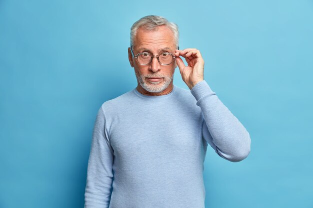 Die Studioaufnahme eines überraschten reifen Mannes sieht mit Staunen aus und trägt eine optische Brille. Der lässige Pullover hört schockierende Nachrichten, die über der blauen Wand isoliert sind und von einem fantastischen Ereignis beeindruckt sind