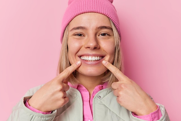 Die süße positive kaukasische Frau zeigt mit dem Zeigefinger auf das Zahnlächeln und zeigt, dass ihre perfekten Zähne Hut und Jacke tragen, die in guter Laune sind, isoliert über rosa Hintergrund Menschen- und Glückskonzept