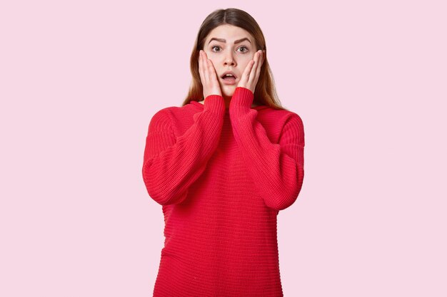 Die Taille einer überraschten jungen Frau berührt die Wangen, trägt einen roten Pullover, drückt einen Schock aus und posiert über rosig