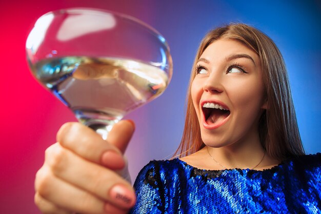 Die überraschte junge Frau in Partykleidung posiert mit einem Glas Wein. Emotionales weibliches süßes Gesicht. Blick aus dem Glas