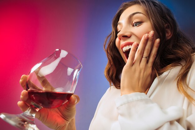 Die überraschte junge Frau in Partykleidung posiert mit einem Glas Wein. Emotionales weibliches süßes Gesicht. Blick aus dem Glas