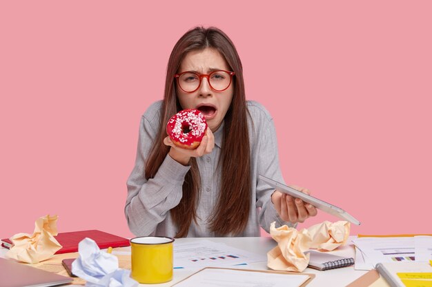 Die unglückliche attraktive junge Frau weint, hat Probleme bei der Arbeit, isst leckeren süßen Donut, trägt ein modernes Touchpad, entwickelt eine Geschäftsstrategie und studiert Grafiken