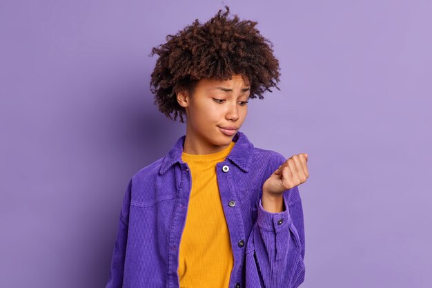 Die unglückliche junge afroamerikanische Frau schaut auf ihre Nägel und möchte eine neue Maniküre in stilvoller Kleidung herstellen.