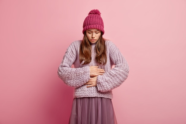 Kostenloses Foto die unglückliche kaukasische frau hält beide hände am bauch, aß verdorbenes essen, hat ein unangenehmes gefühl im bauch, trägt eine rosa kopfbedeckung mit pompon, einen strickpullover und einen faltenrock und steht über einer rosa wand